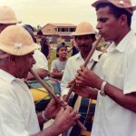 Fotografia colorida. Destaque para dois homens à frente de um grupo de pessoas. Eles estão tocando um instrumento de sopro, cujo formato é cilíndrico, estreito e comprido. Ambos vestem camisas sociais brancas e chapéu marrom. O homem à esquerda possui cabelo grisalho e baixa estatura, o homem à direita é mais jovem e de maior estatura. As pessoas ao fundo observam a cena, dois homens vestem trajes semelhantes aos dois anteriores e carregam instrumentos percussivos.
