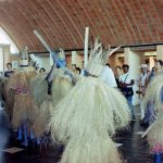 Fotografia colorida. Em uma área coberta por laje de tijolos, um grupo de pessoas com roupas à caráter está sendo assistido por outras pessoas ao fundo. Eles apresentam a pele pintada de azul, vestem túnicas de palha e utilizam um adorno de penas sobre suas cabeças. O homem à direita na imagem e um homem ao centro carregam, cada um, um bastão estreito e comprido em suas mãos.