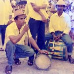 Fotografia colorida. À esquerda, um homem agachado, apoiando um instrumento de sopro em um dos joelhos flexionados, com uma mão sobre um tambor ao seu lado. Ele olha para frente. Ao seu lado, outro homem está sentado sobre um grande tambor, segurando uma baqueta para bumbo. Ao redor deles há muitas pessoas e dois homens em pé, segurando flautas semelhantes à do primeiro homem descrito.