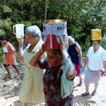 Fotografia colorida. Um grupo de mulheres caminhando em uma estrada pedregosa, margeada por arbustos. Duas delas carregam latas sobre suas cabeças, outra carrega um jarro branco erguido à altura de seu ombro. Um menino carrega uma pequena lata metálica e as acompanha.